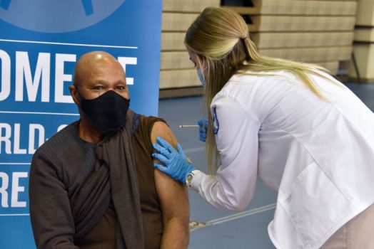 woman in white coat giving a shot to person sitting in a chair, masked