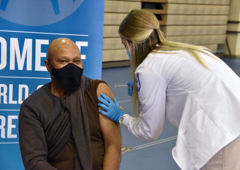 woman in white coat giving a shot to person sitting in a chair, masked