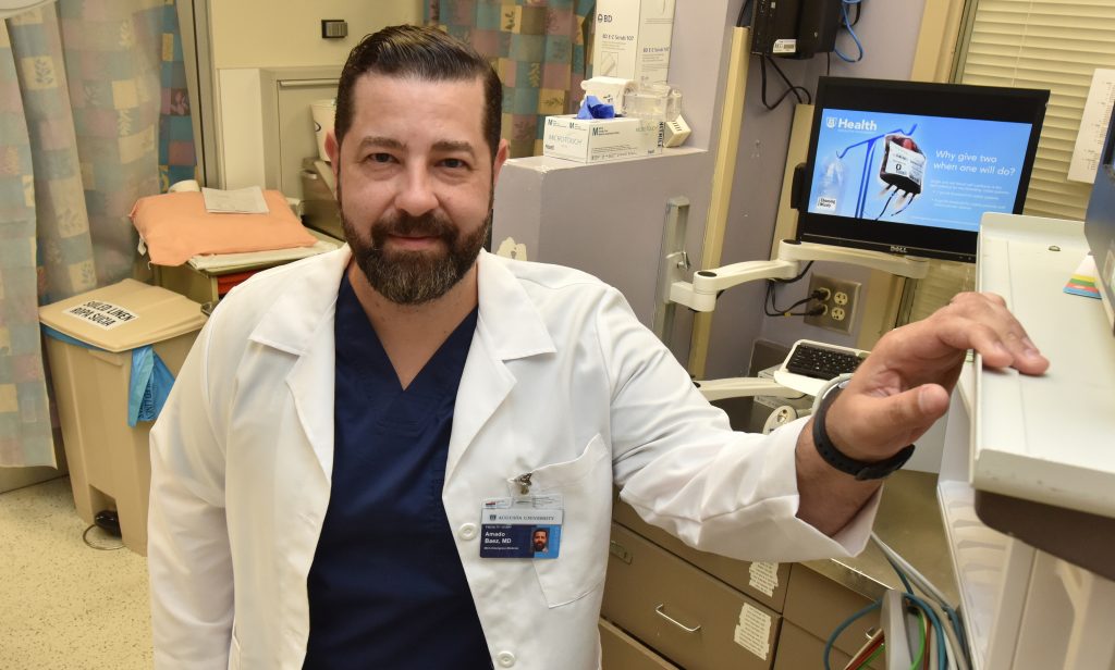 Man with brown hair and beard, in white coat, stands in emergency room