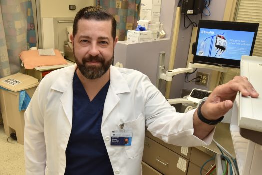 Man with brown hair and beard, in white coat, stands in emergency room