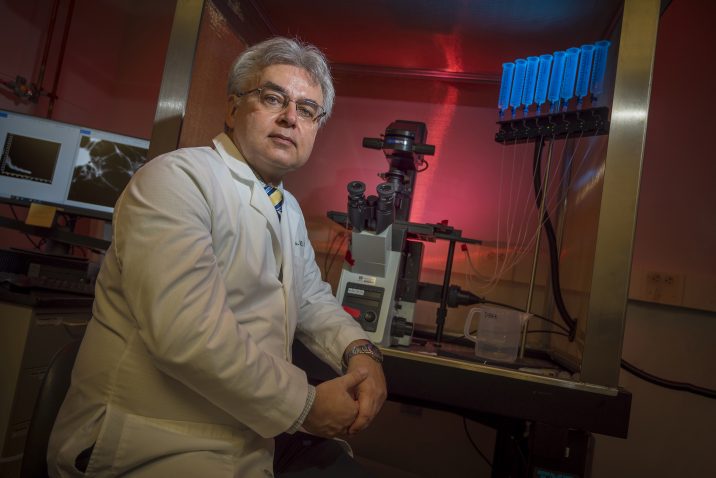 Gray haired man in white coat sits at lab bench with microscope in background
