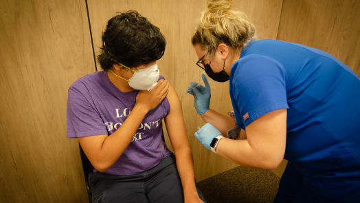 a masked woman administers a vaccine to a masked man