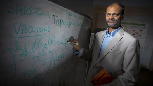 Man in cream jacket and blue shirt writes math equations on white board