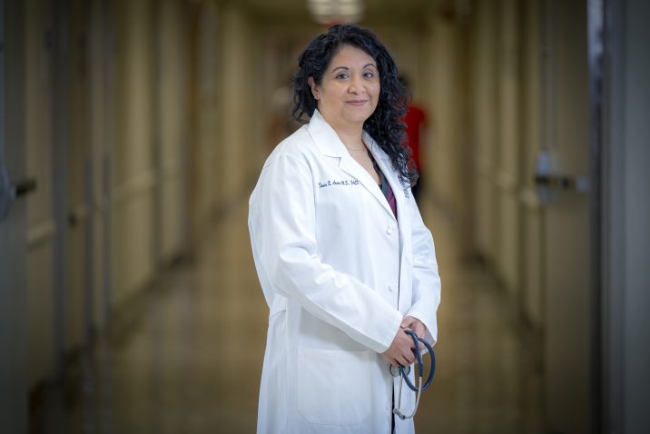 A woman with curly dark hair, wearing a white coat, stands in a hallway looking at the camera