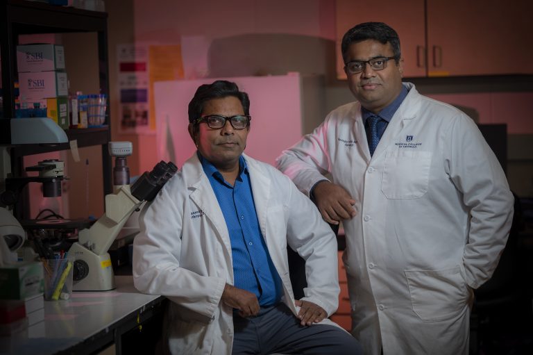 Two men with dark hair and wearing glasses and white coats stand side by side in lab