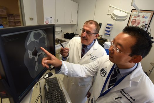 Two men with dark hair in white jackets, pointing at screen with skull xray