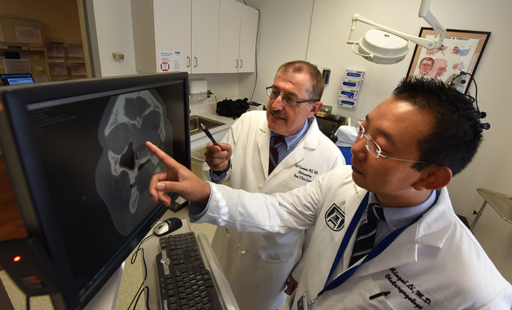 Two men with dark hair in white jackets, pointing at screen with skull xray