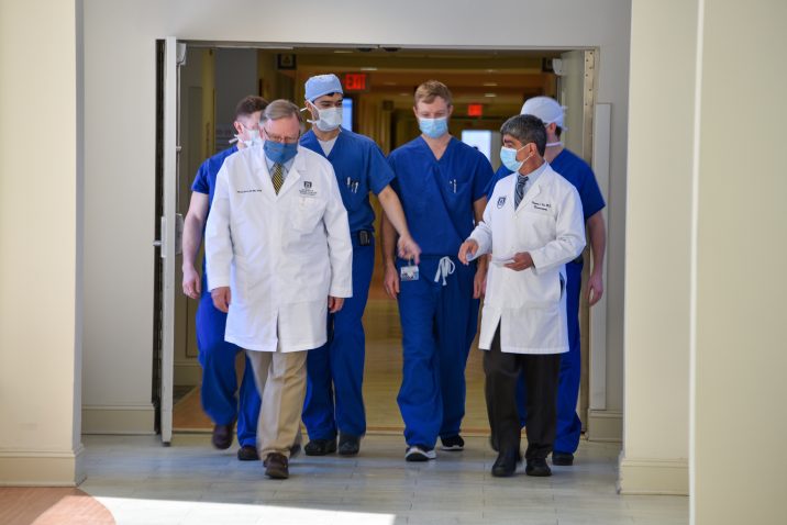 Two doctors in white coats lead a group of younger doctors in blue scrubs through a hospital hallway