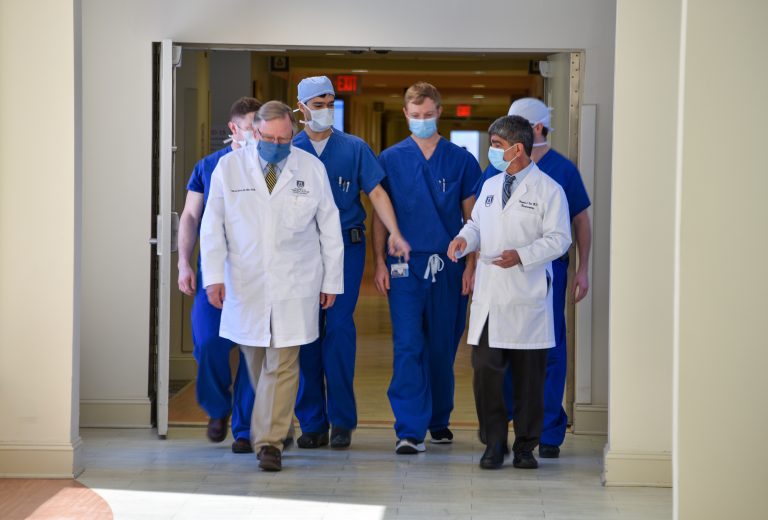 Two doctors in white coats lead a group of younger doctors in blue scrubs through a hospital hallway
