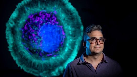 Man with glasses looks at camera with image of cell behind him