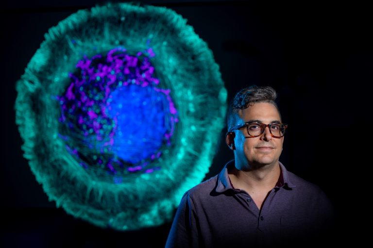Man with glasses looks at camera with image of cell behind him