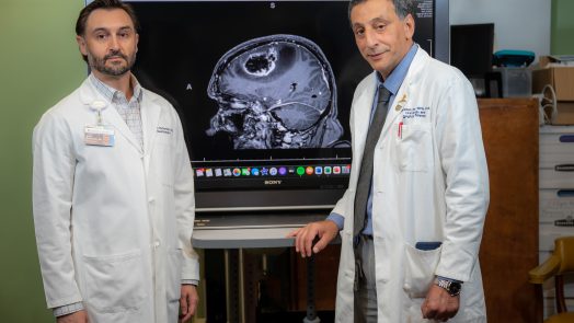 Two men in white coats stand in front of a scan showing a brain tumor