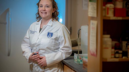 Woman with curly hair, wearing white coat stands in lab