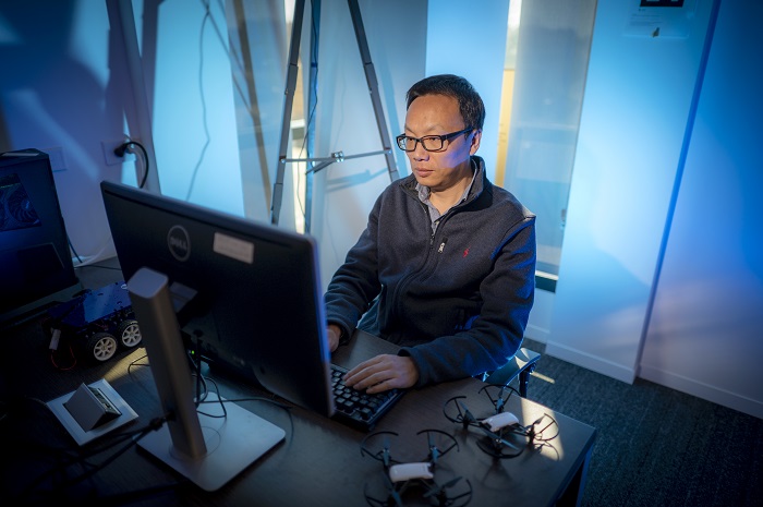 man with glasses sitting at computer with quadcopter beside him