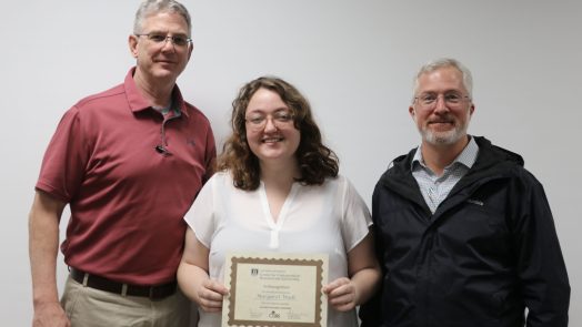 a woman stands between two men, holding a certificate