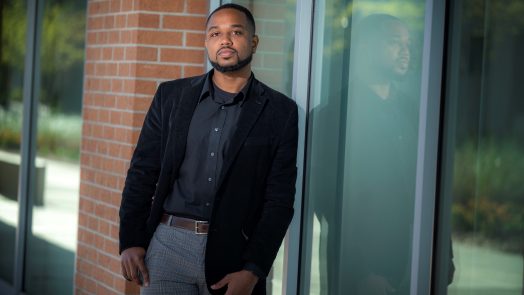 Man in black coat and shirt leans against building