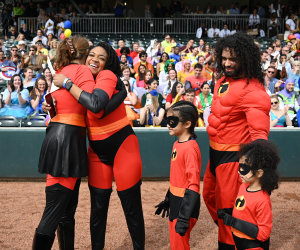 two women hug in celebration as two kids and one man look on, all dressed as superheroes