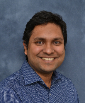 A man in a blue printed polo shirt smiles at the camera in front of a plain blue background