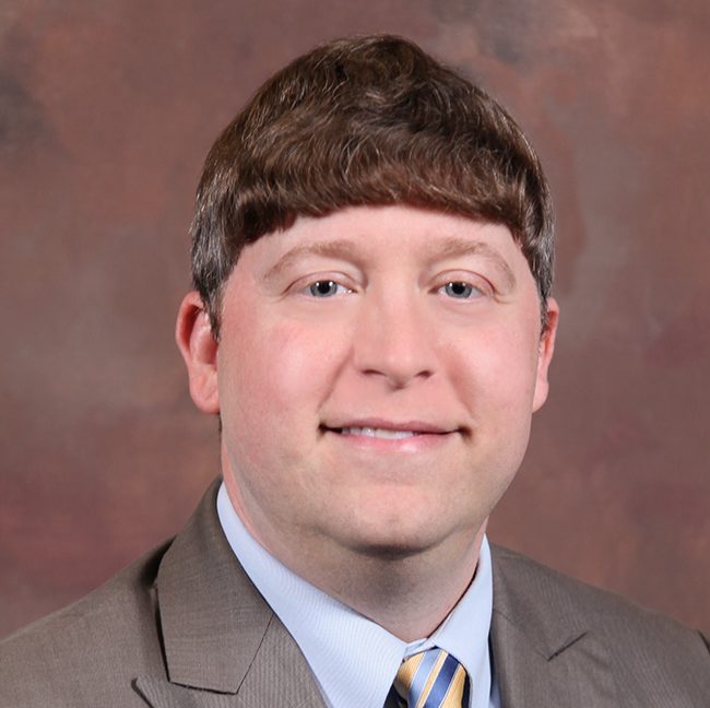 A man in a brown suit jacket, light blue shirt and gold and blue tie smiles at the camera