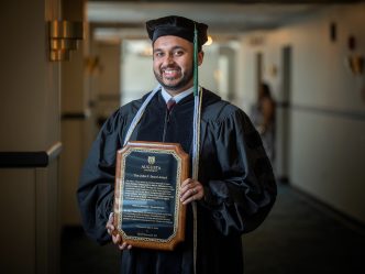 Man stands in graduation robe and cap holding plaque