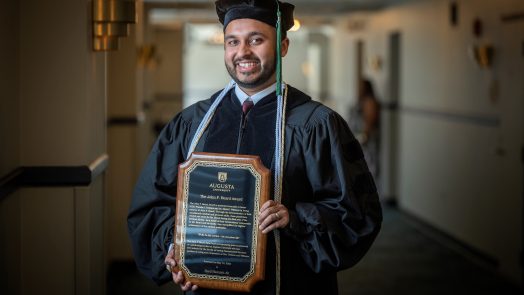 Man stands in graduation robe and cap holding plaque