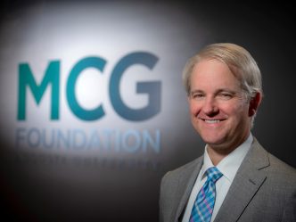 Man in gray suit stands in front of sign that says MCG Foundation