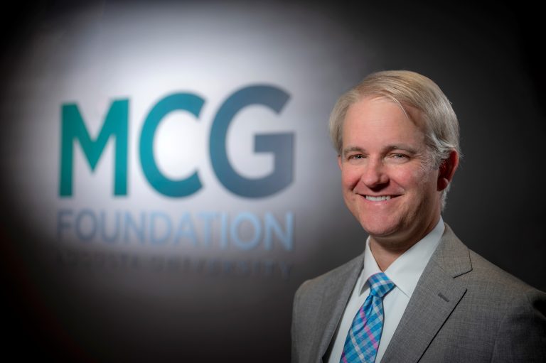 Man in gray suit stands in front of sign that says MCG Foundation