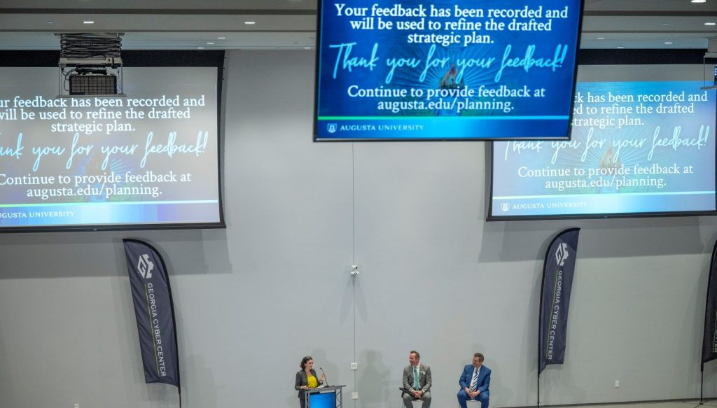 woman stands at podium; above her, digital signs encouraging readers to submit a feedback survey