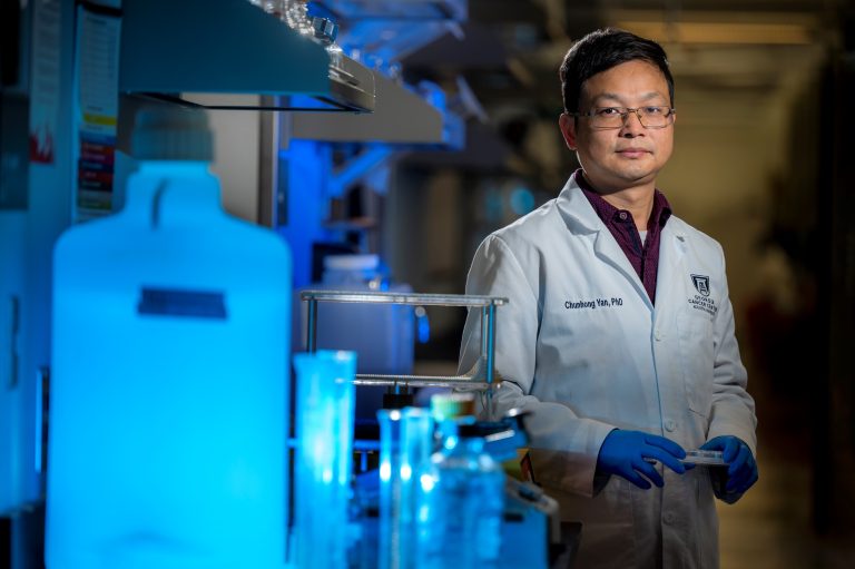 Man in a scientific lab coat and glasses stands in front of beakers in lab