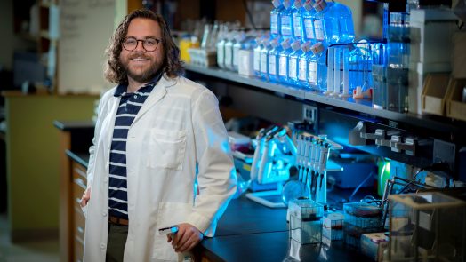 Man with medium length brown curly hair and glasses stands in white coat in lab