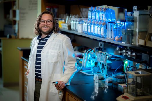 Man with medium length brown curly hair and glasses stands in white coat in lab