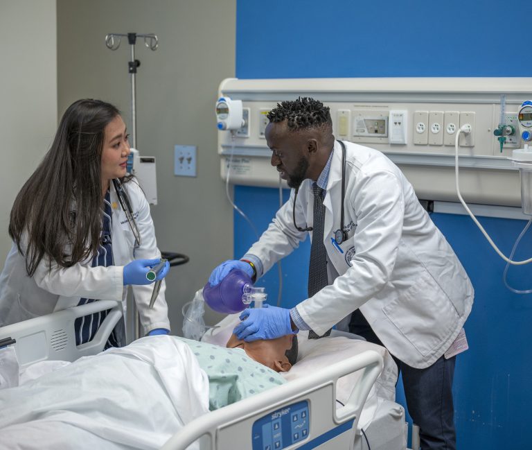Male and female medical students in white coats stand over mannequin in hospital like room