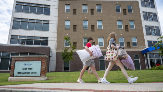 two women carrying bags walk by the front of a residence hall