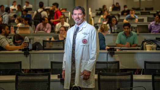 Man in white coat stands in front of classroom of students