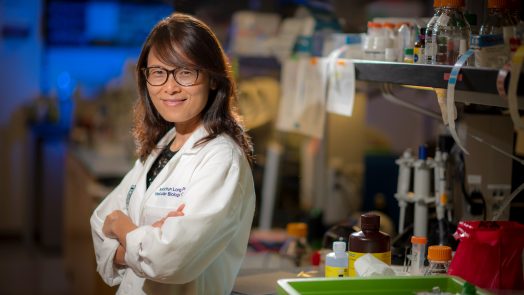 Woman with brown hair and glasses in white coat leans against lab bench