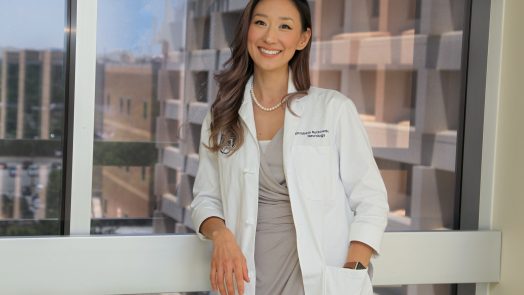 Woman with long brown hair in white coat and gray dress leans against window