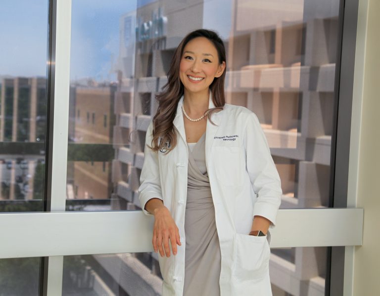 Woman with long brown hair in white coat and gray dress leans against window