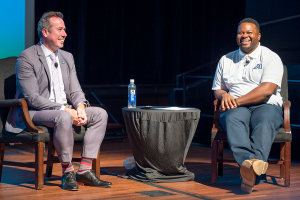 two men sit in chairs talking and smiling