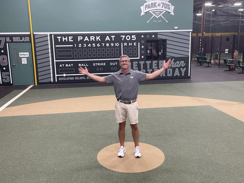 Man stands with his arms raised on an indoor baseball field.