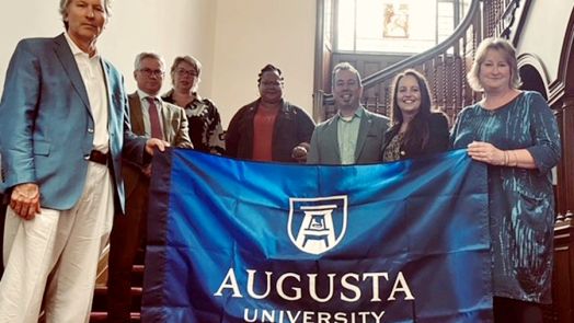 seven people stand on stairs hold up a blue flag with the Augusta University logo in white