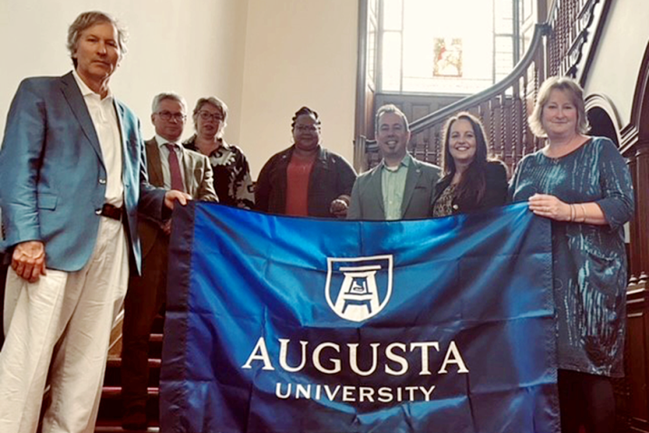 seven people stand on stairs hold up a blue flag with the Augusta University logo in white