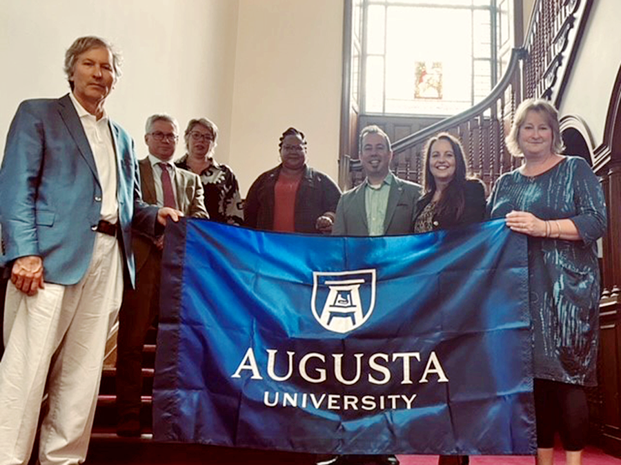 seven people stand on stairs hold up a blue flag with the Augusta University logo in white