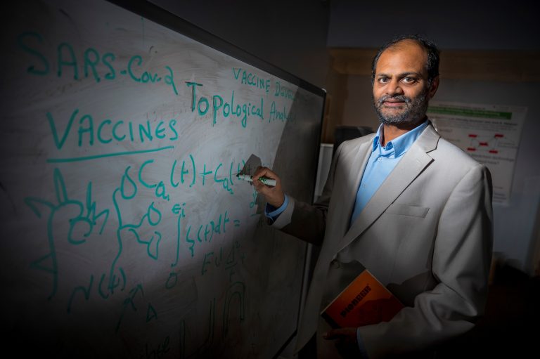 Man in gray jacket stands in front of white board with marker in hand