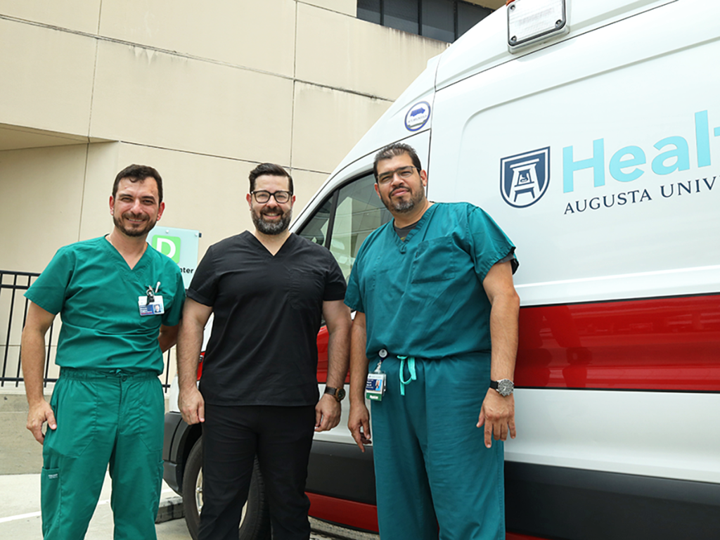 Three male doctors wearing scrubs stand in front of an ambulance