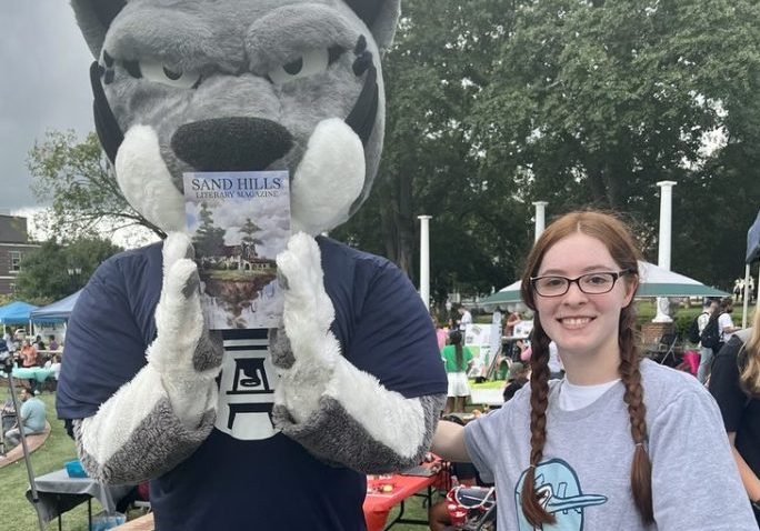 Augustus the mascot holding the new edition of Sand Hills Literary Magazine. A student is standing next to him while hugging him.