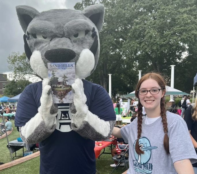 Augustus the mascot holding the new edition of Sand Hills Literary Magazine. A student is standing next to him while hugging him.