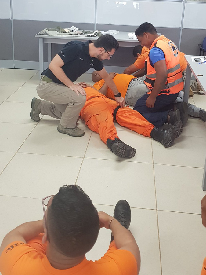 Three men practice checking vital signs of patients for emergency procedures in a classroom as others watch.