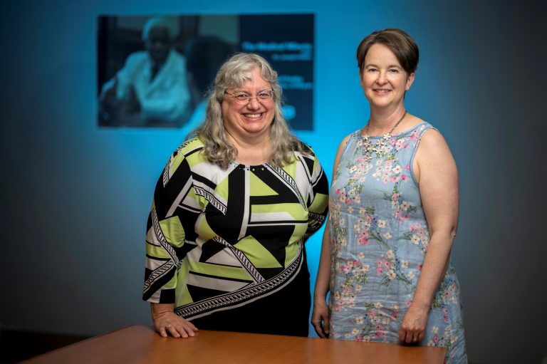 Two women stand in front of poster smiling at camera