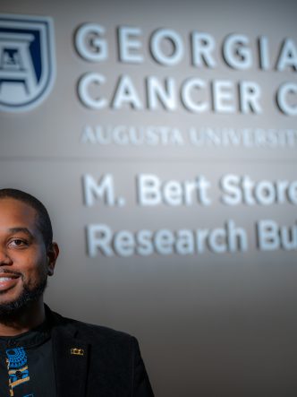 Man in black coat and blue and black shirt stands in front of sign that reads "Georgia Cancer Center"