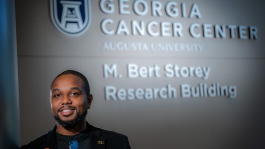 Man in black coat and blue and black shirt stands in front of sign that reads "Georgia Cancer Center"